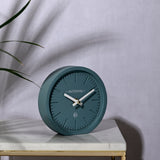 Minimalist round matt desk and mantel clock, 15.4cm, in teal with baton hour markers and white hands, shown on a marble table beside a plant.