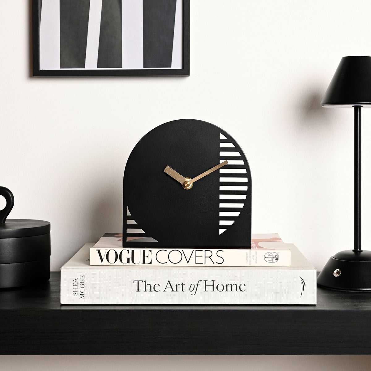Black arched steel mantel clock with gold hands and cut-out horizontal stripe detailing, displayed on stacked books on a dark shelf.