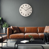 Round metal wall clock with white dial, black Arabic numerals and slim black rim, shown above a tan leather sofa on a dark grey living room wall.