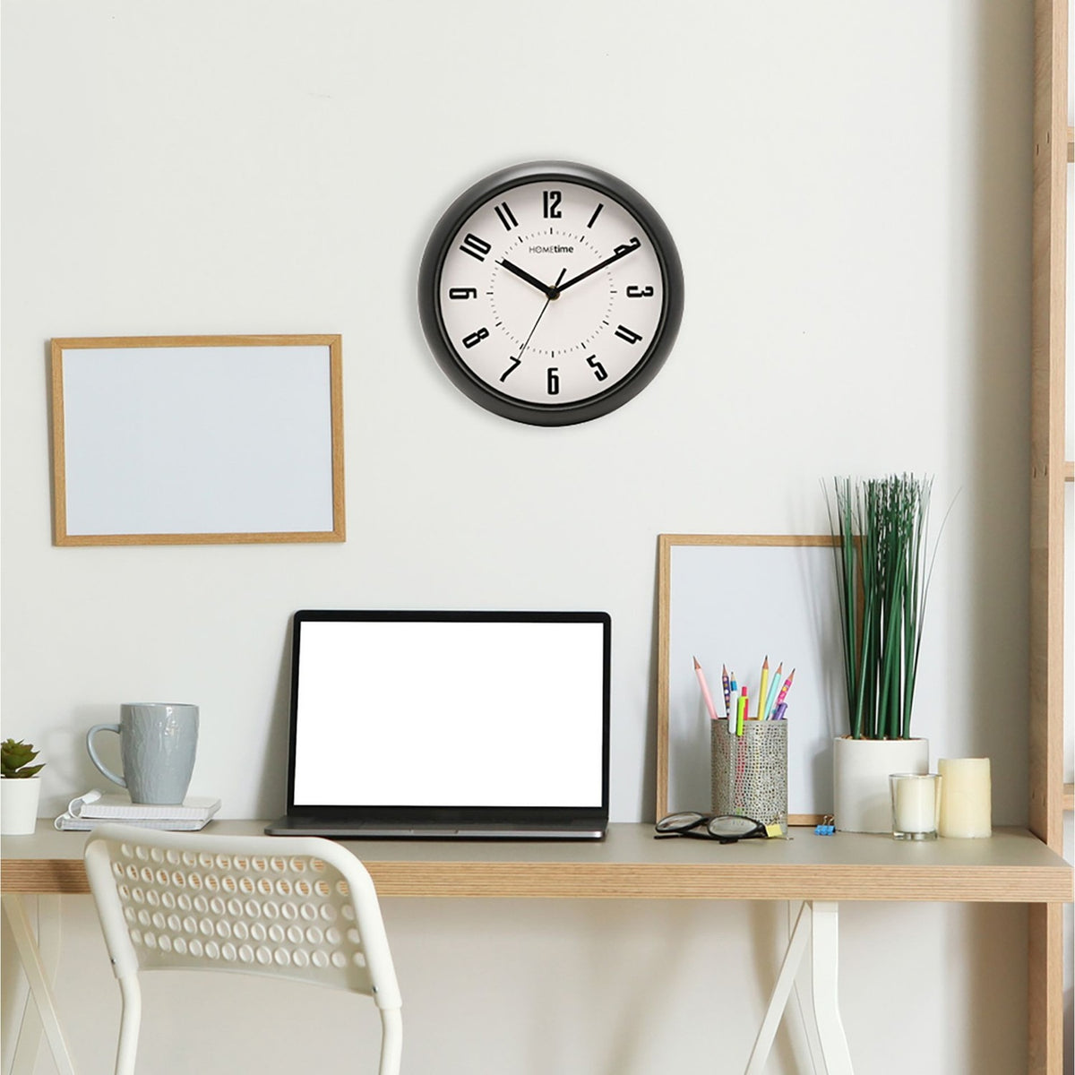 Round wall clock with black frame and white dial featuring bold numerals, displayed on a wall above a home office desk setup.