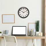 Round wall clock with black frame and white dial featuring bold numerals, displayed on a wall above a home office desk setup.
