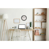 Round wall clock, 25.5cm, with black frame and white dial featuring bold numerals, displayed above a home office desk with laptop and shelving.