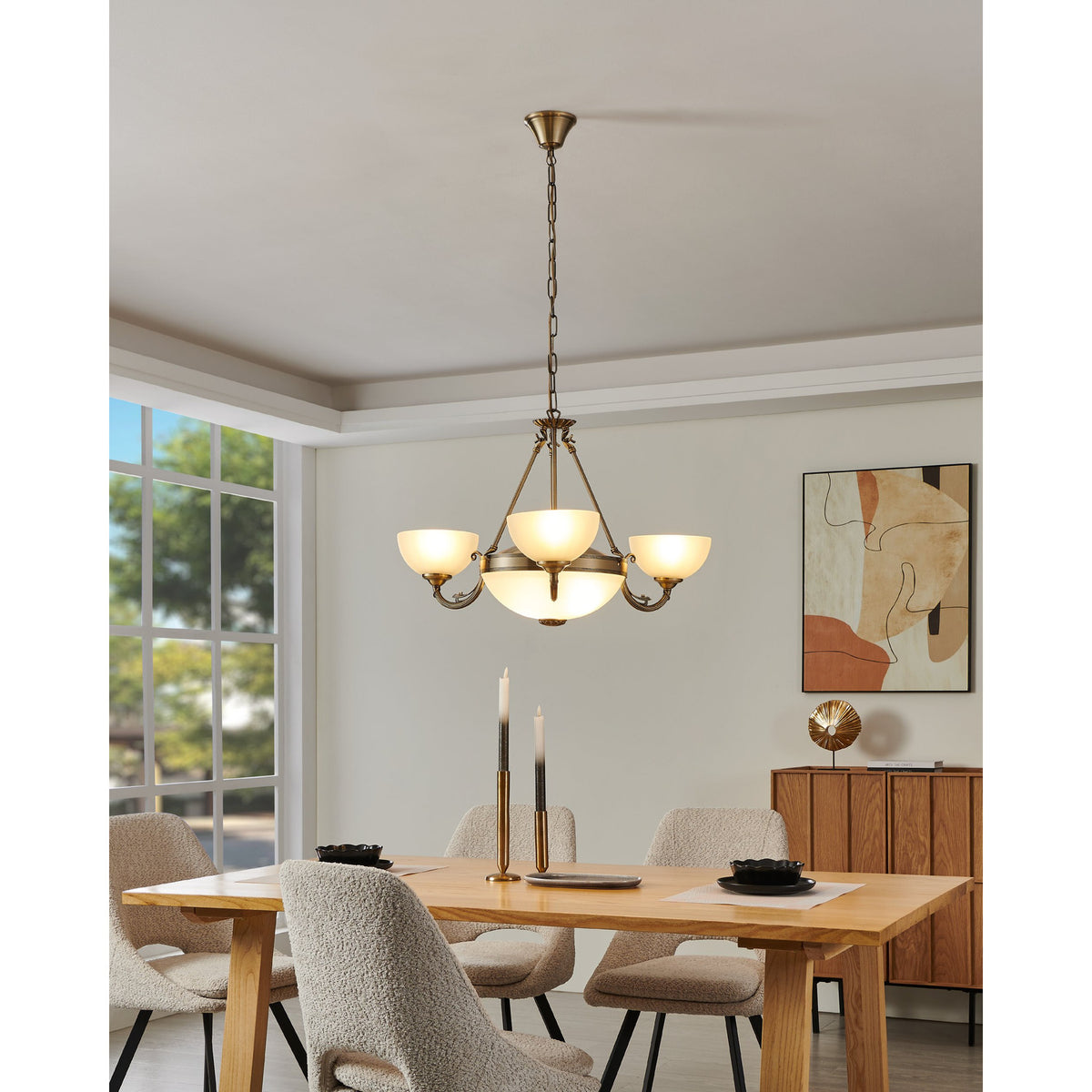Walbane Glass 3 Light Pendant in bronze, with scrolled metal arms and upturned white satin glass shades around a central frosted glass bowl, hung above a dining table.
