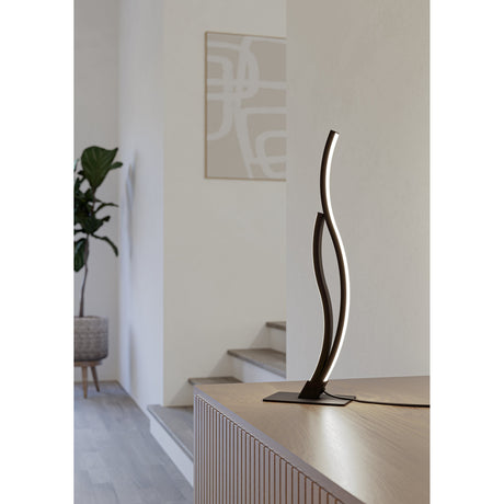 Wasmane LED Table Lamp in black, with two wavy steel arms rising from a flat base, shown lit on a wooden sideboard in a hallway setting.