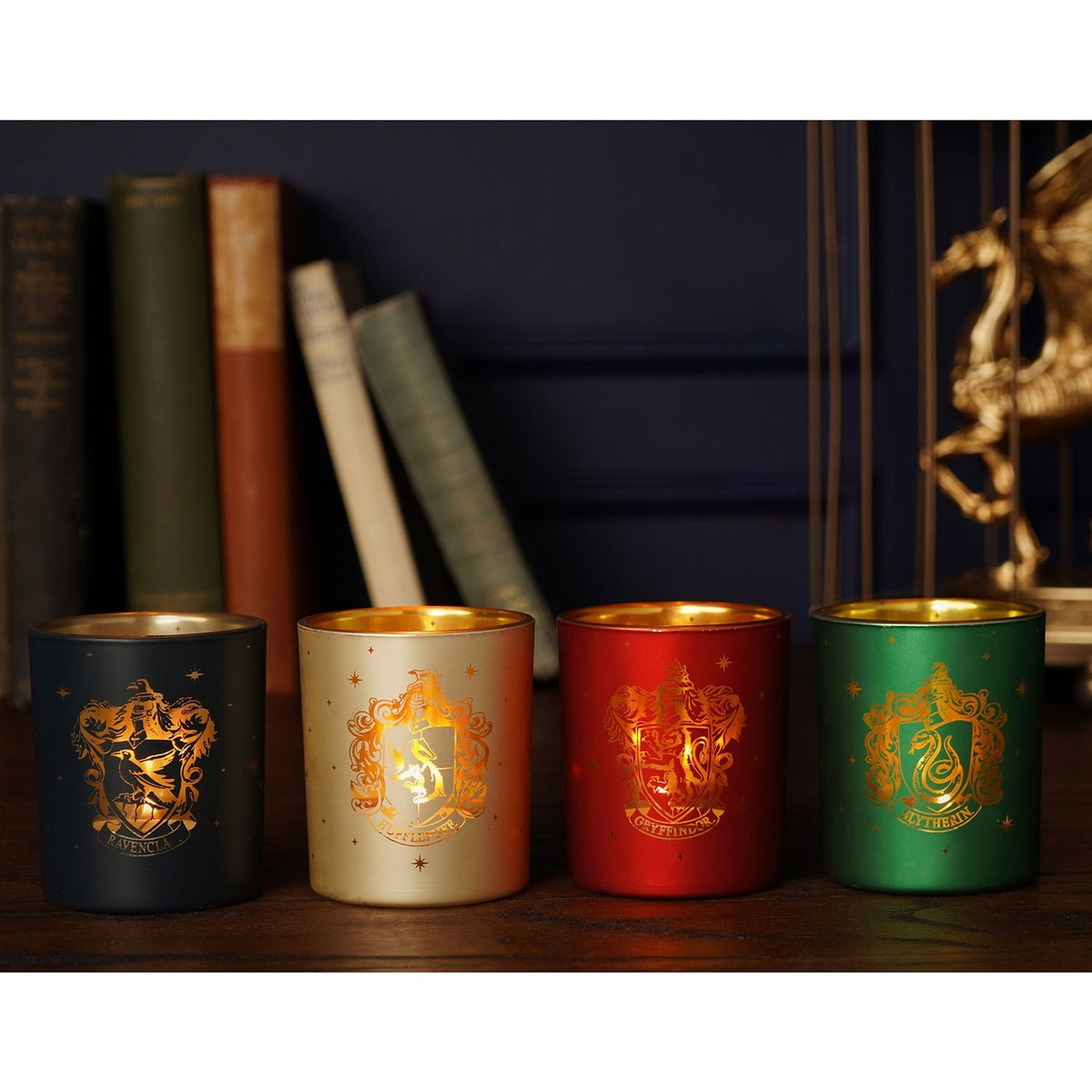 Set of 4 round glass tealight candle holders in black, cream, red and green, each with a gold Hogwarts house crest, displayed before books and a gold dragon figure.