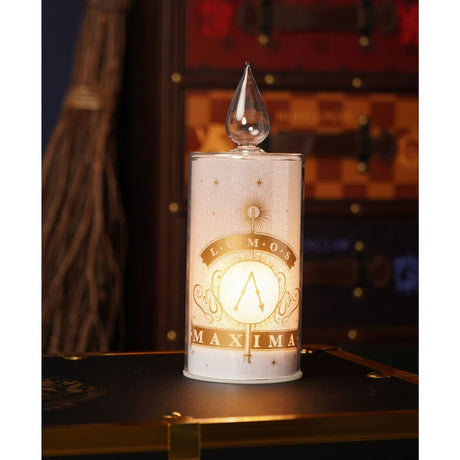 Harry Potter Lumos Maxima Glass LED Candle, 22cm white pillar with gold wand and starburst design, clear teardrop flame top, lit on a dark surface with trunk and broomstick in background.