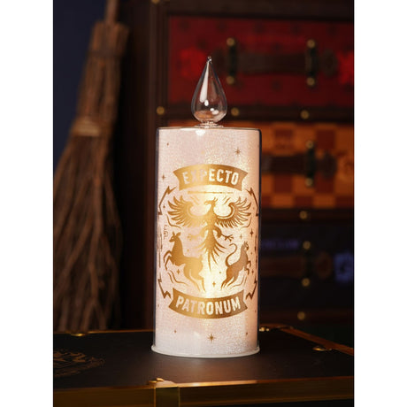 Harry Potter Patronus Crest Glass LED Candle, 26cm white pillar with gold "Expecto Patronum" design and clear flame tip, lit on a dark surface before a trunk.