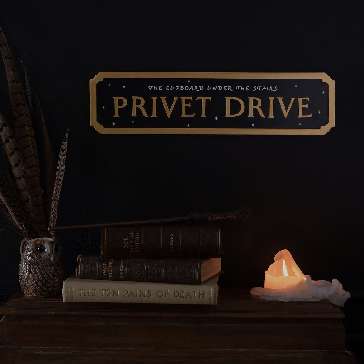 Black rectangular "Privet Drive" street sign wall plaque with gold raised lettering, notched gold border, and star details, displayed above books, a wand, and a candle.