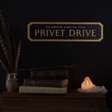 Black rectangular "Privet Drive" street sign wall plaque with gold raised lettering, notched gold border, and star details, displayed above books, a wand, and a candle.