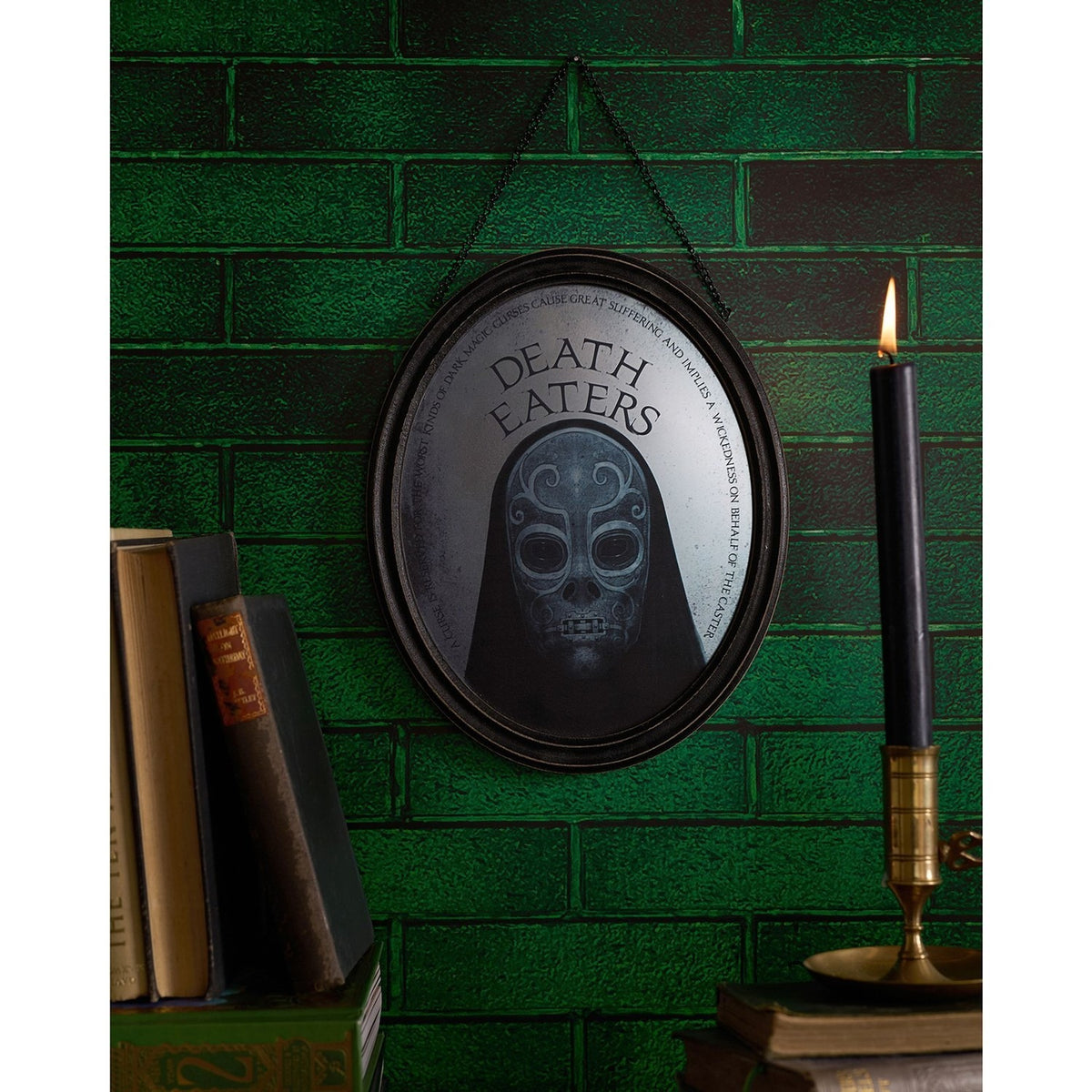 Harry Potter Death Eater oval mirrored wooden plaque in a black frame, hooded mask design with "DEATH EATERS" text, hung on a green brick wall beside books and a candle.