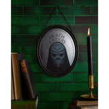 Harry Potter Death Eater oval mirrored wooden plaque in a black frame, hooded mask design with "DEATH EATERS" text, hung on a green brick wall beside books and a candle.