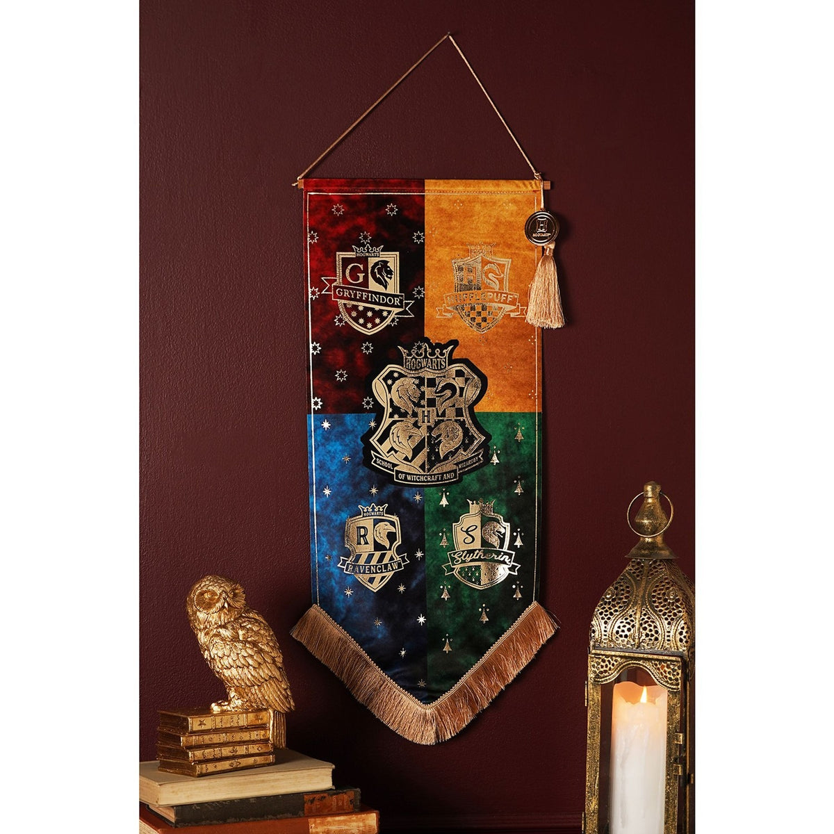 Harry Potter Hogwarts Houses fringed wall banner, pointed pennant quartered in red, orange, blue and green with gold house crests and central Hogwarts shield, tassel detail, hung on a dark red wall.