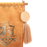 Close-up of a Harry Potter Hogwarts fringed wall banner with a gold Hufflepuff crest on mustard fabric, wooden rail, round Hogwarts medallion charm, and tassel, on a white background.