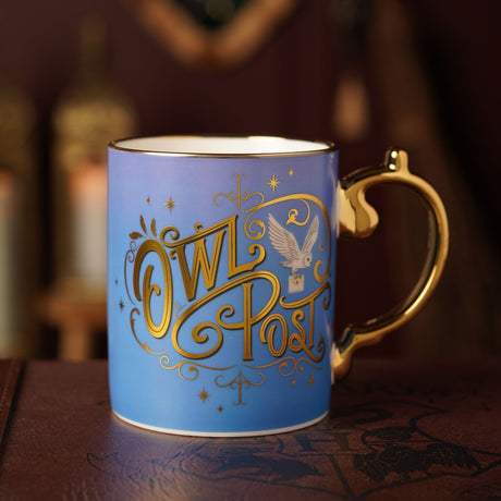 Porcelain mug in blue with gold "Owl Post" script, white owl illustration, gold rim band and curled gold handle, shown on a dark wooden surface.