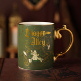 Green porcelain Diagon Alley mug with gold handle, featuring gold Gothic lettering, scrollwork, shopfronts, a domed building, and owl motifs.