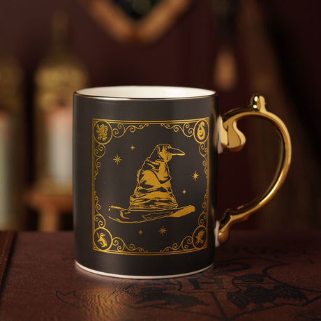 Porcelain mug with matt black body and gold Sorting Hat design, scrolling border, star details and house emblems, gold handle, shown on a dark surface.