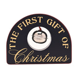 Arched black wooden plaque reading "THE FIRST GIFT OF Christmas" in gold lettering, with a silver-coloured sleigh bell set in a round opening, on a plain white background.