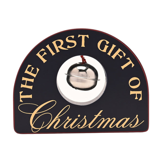 Arched black wooden plaque reading "THE FIRST GIFT OF Christmas" in gold lettering, with a silver-coloured sleigh bell set in a round opening, on a plain white background.