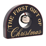 Arched black wooden plaque reading "THE FIRST GIFT OF Christmas" in gold lettering, with a round cutout holding a silver-coloured bell, shown on a white background.