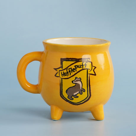 Yellow stoneware mug shaped like a cauldron with three short feet, featuring the Hufflepuff crest and badger design, set against a plain light blue background.