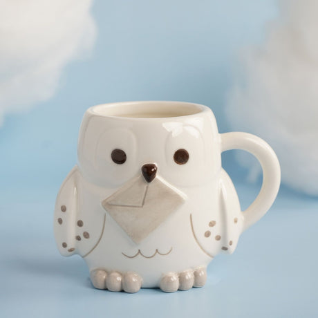 White ceramic stoneware mug shaped as Hedwig the owl, with round dark eyes, a brown beak, a grey envelope on her chest, and speckled wings, set against a light blue background.