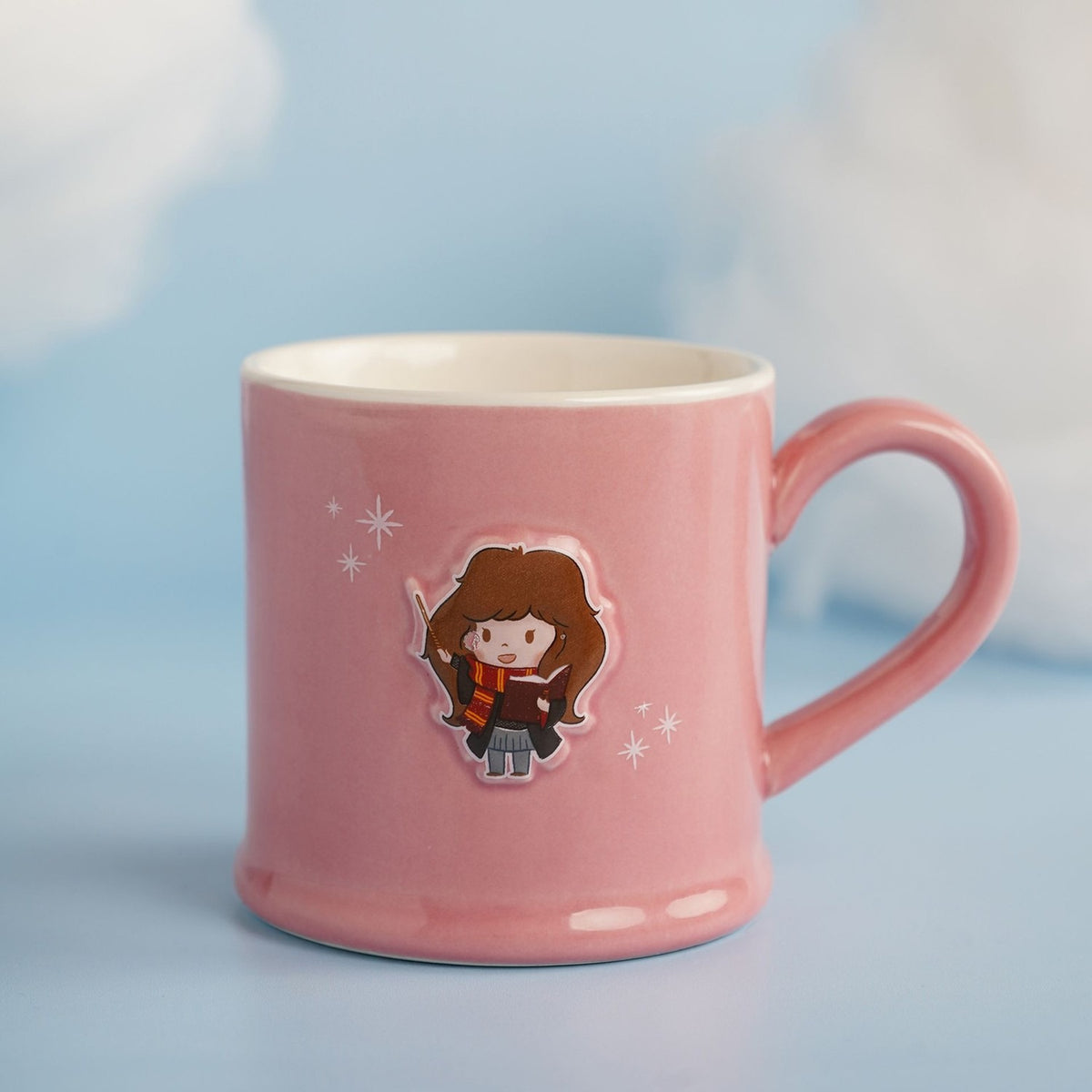 Pink stoneware mug with an embossed cartoon Hermione Granger design holding a wand and book, white interior, pink handle, set against a soft blue background.