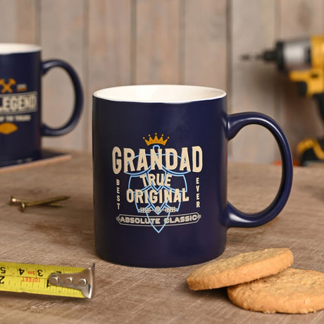 Blue stoneware mug with white interior, printed "GRANDAD TRUE ORIGINAL ABSOLUTE CLASSIC" design in cream and gold, shown on a wooden table beside another mug, biscuits, and tools.