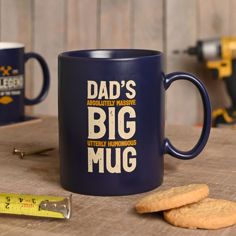 Blue stoneware pint mug, 580ml, with "Dad's Absolutely Massive Big Utterly Humongous Mug" in cream and amber text, shown on a wooden table beside biscuits and tools.
