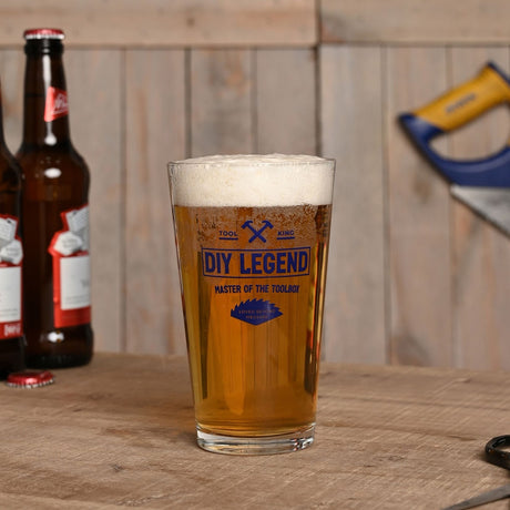 Clear tapered pint beer glass, 460ml, filled with beer, printed with navy "DIY Legend, Master of the Toolbox" design, displayed on a wooden table beside beer bottles.