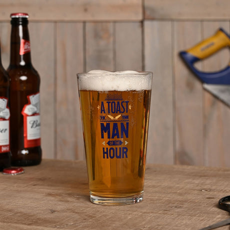 Clear tapered pint beer glass, 460ml, filled with beer, printed with "A Toast To The Man of The Hour" in blue vintage-style lettering, shown on a wooden table with beer bottles.