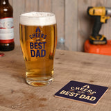 Clear pint beer glass filled with beer, printed with "Cheers To The Best Dad" in navy blue, beside a matching navy coaster on a wooden table.