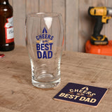 Clear pint beer glass with navy blue "Cheers to the Best Dad" text and matching coaster, shown on a wooden table with a beer bottle and drill in the background.