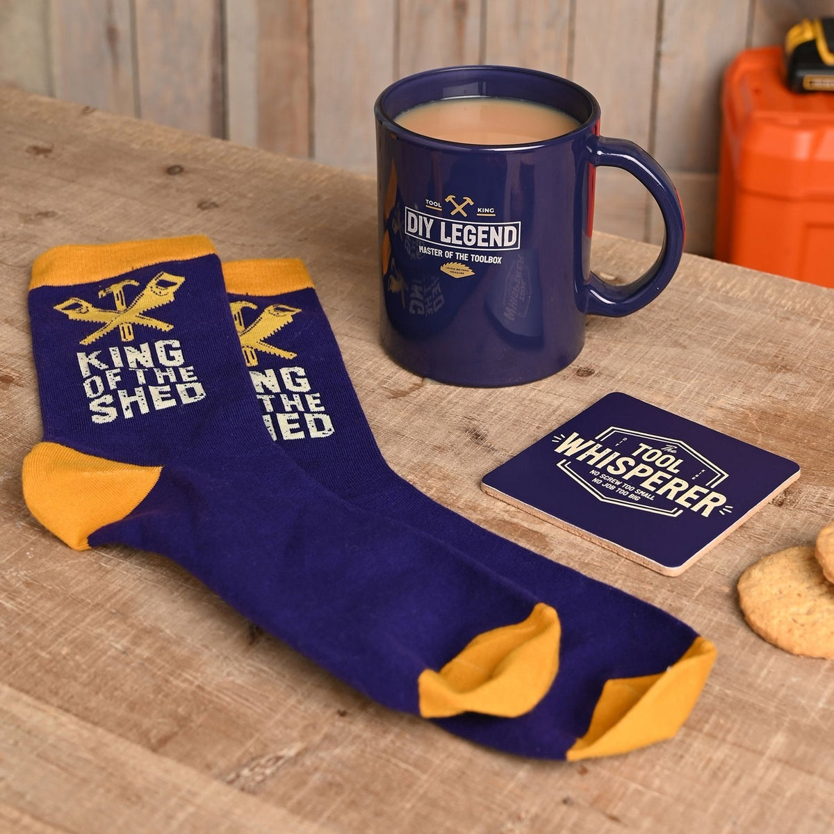 DIY Legend Mug gift set with tea, shown alongside blue cotton socks with mustard cuffs and a matching square coaster reading Tool Whisperer, on a wooden surface.