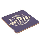 Square blue coaster with gold "The Tool Whisperer" text, cork backing, shown tilted on a white background.
