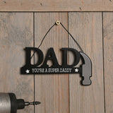 Wooden DAD wall plaque in black with "You're A Super Daddy" text and white stars, the last letter shaped as a hammer with silver head, hung on a wooden panel.