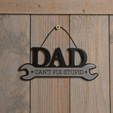 Wooden spanner-shaped wall plaque reading "DAD CAN'T FIX STUPID" in black and white, hung on a rustic wooden plank wall.