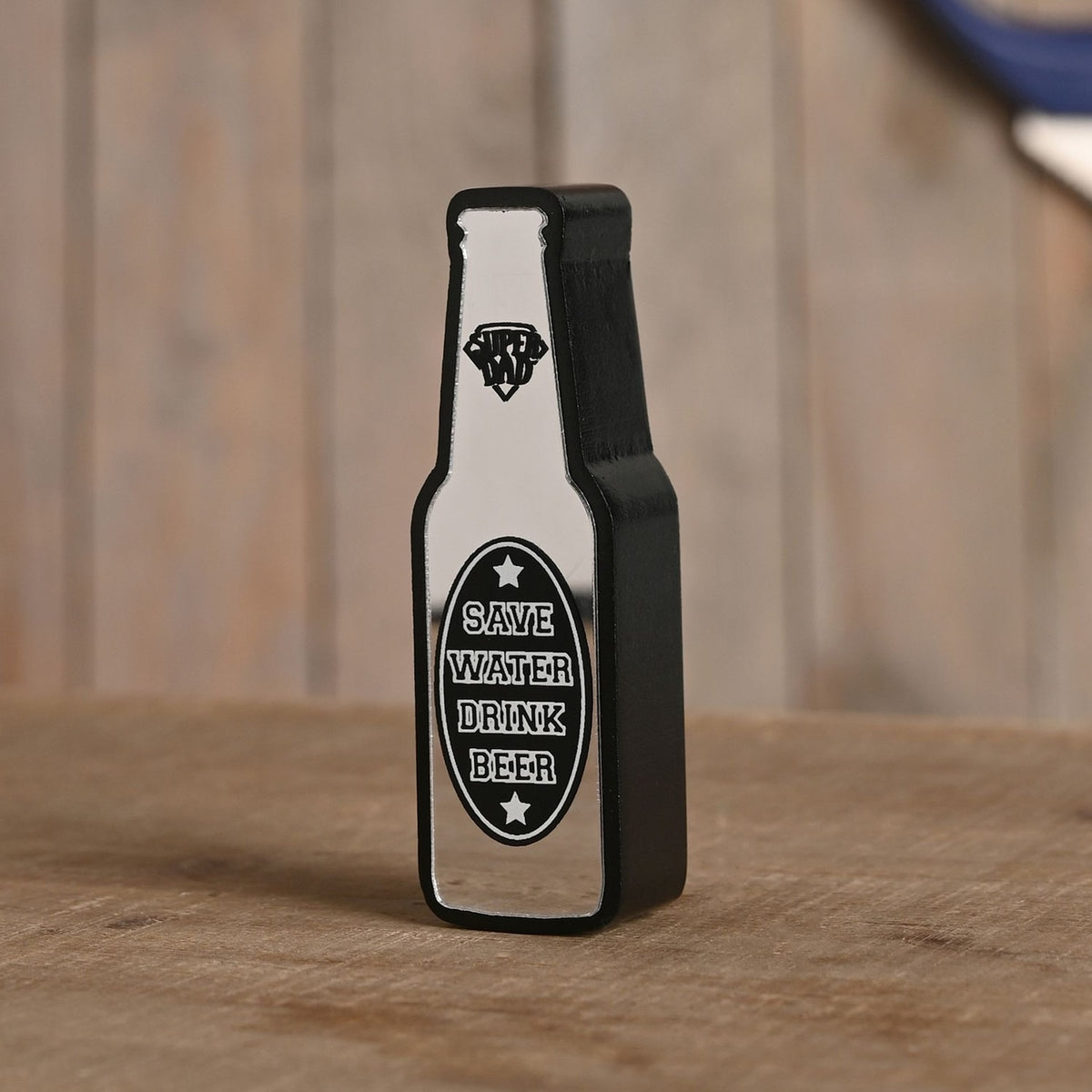 Wooden bottle-shaped wall plaque with mirrored silver finish and black edge, black oval label reading "Save Water Drink Beer" in white, standing on a wooden surface.