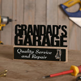 Wooden standing plaque with black cut-out letters reading Grandad's Garage, black panel reading Quality Service and Repair with a small silver tool charm, displayed on a workbench with tools.