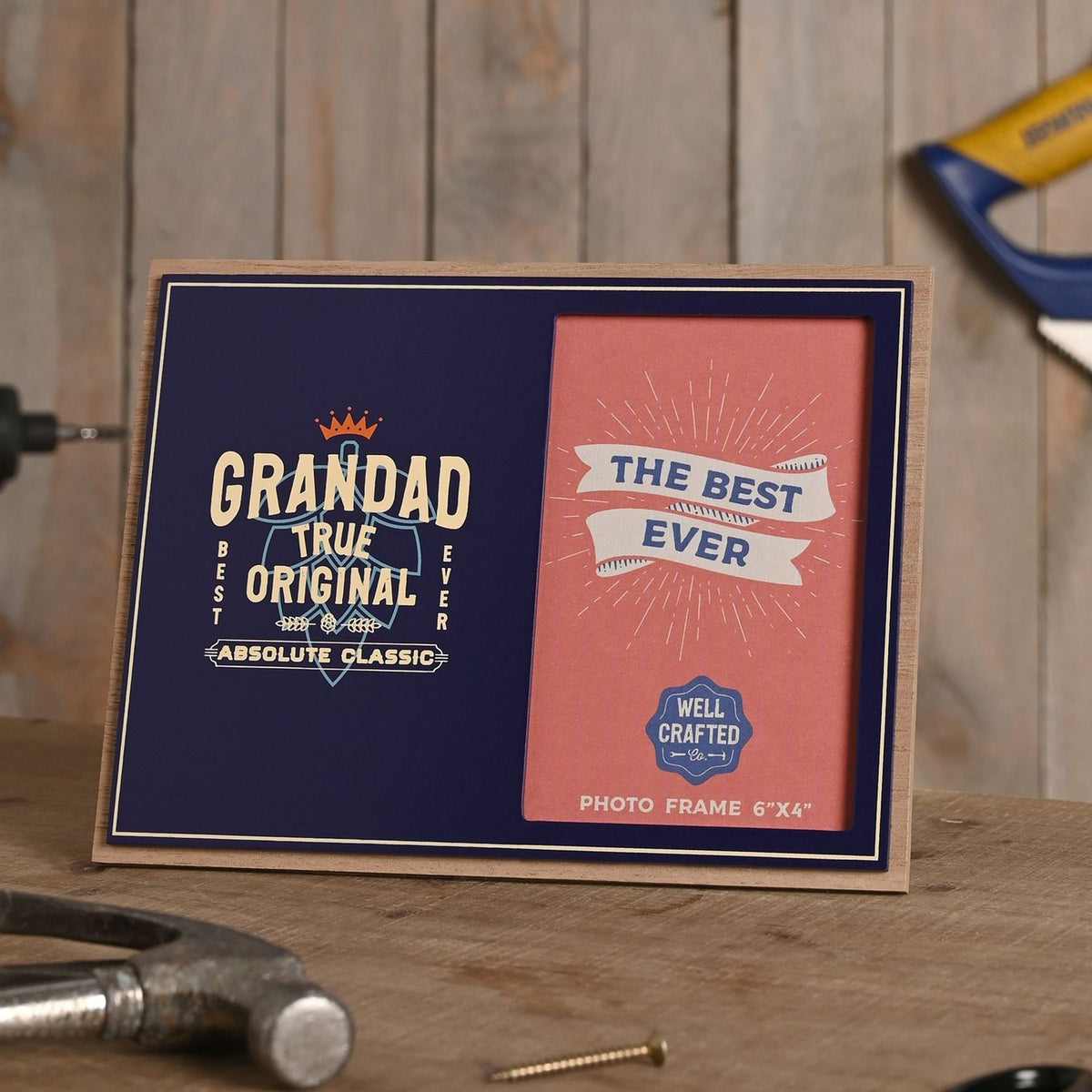 Wooden landscape photo frame, 6x4in, with navy blue front reading "Grandad True Original, Absolute Classic", crown detail, beside a pink "The Best Ever" photo insert, shown on a workbench with tools.