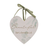 White wooden heart-shaped hanging plaque with pale green leaf print, "Beautiful bridesmaid" in gold script and typewriter lettering, hung by a sage green ribbon bow, on a plain white background.