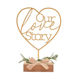 Gold wire heart mantel plaque reading "Our love Story" on a slim stem, set in a wooden block base with artificial leaves and a jute bow, on a plain white background.