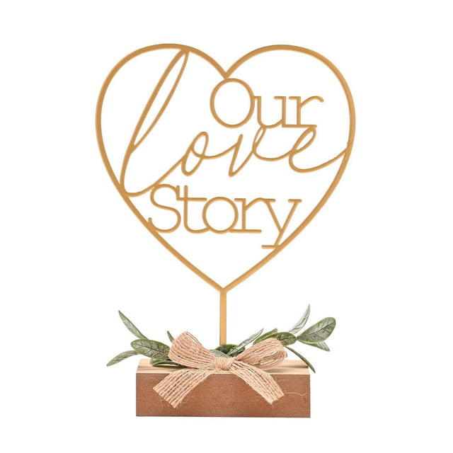Gold wire heart mantel plaque reading "Our love Story" on a slim stem, set in a wooden block base with artificial leaves and a jute bow, on a plain white background.
