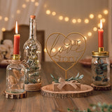 Gold heart mantel plaque reading "Our Love Story" on a slim stem, set in a wooden block base with leaf sprig and bow, displayed on a wood slab amid candles and bottles.
