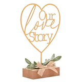 Gold metal heart mantel plaque reading "Our love Story" on a slim stem, set in a wooden block base with green leaf sprig and burlap bow, on a white background.