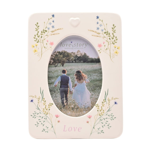 White ceramic photo frame with oval window for a 6 x 4in photo, pastel wildflower sprays at each corner, a raised heart detail, and "Love" printed below in pink.