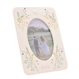 White ceramic photo frame with oval window, pastel wildflower sprays at each corner, raised heart detail, and "Love" printed below, holding a 6 x 4in photo on a plain background.