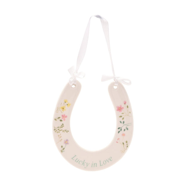 Off-white ceramic horseshoe wall art with painted wildflowers, "Lucky in Love" in green script, hung by a white ribbon bow on a plain white background.