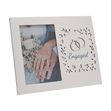 White wooden 6x4in photo frame with a laser cut panel of leaves, hearts and interlinked rings around "Engaged" in blue, beside a bride holding a bouquet.