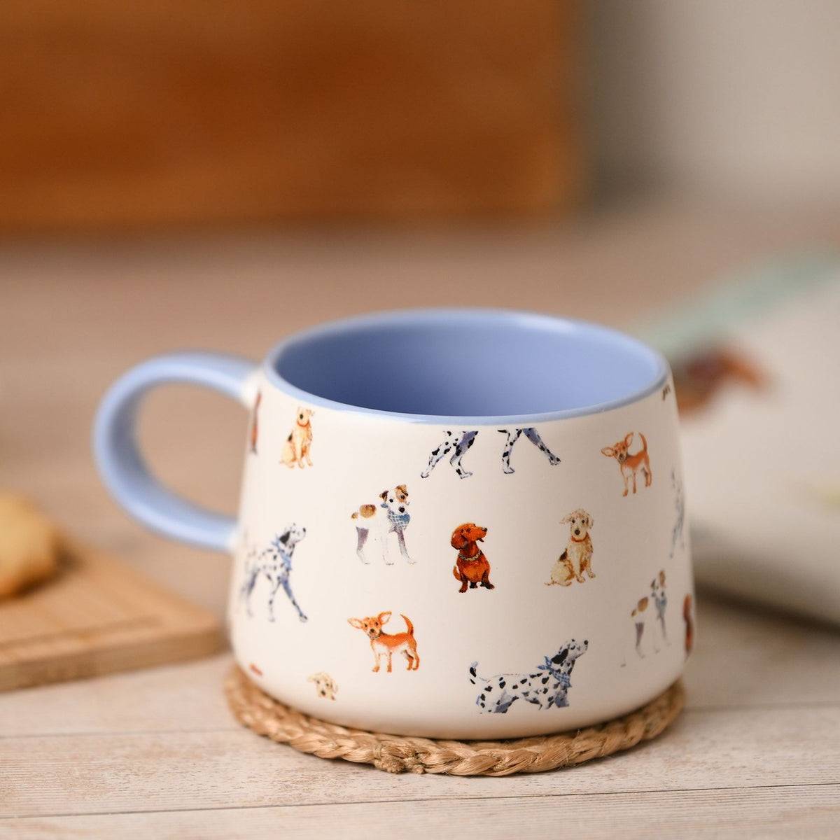 Tapered ceramic mug in white with an all-over watercolour dog pattern, pale blue handle and interior, shown on a woven coaster near a wooden tray.