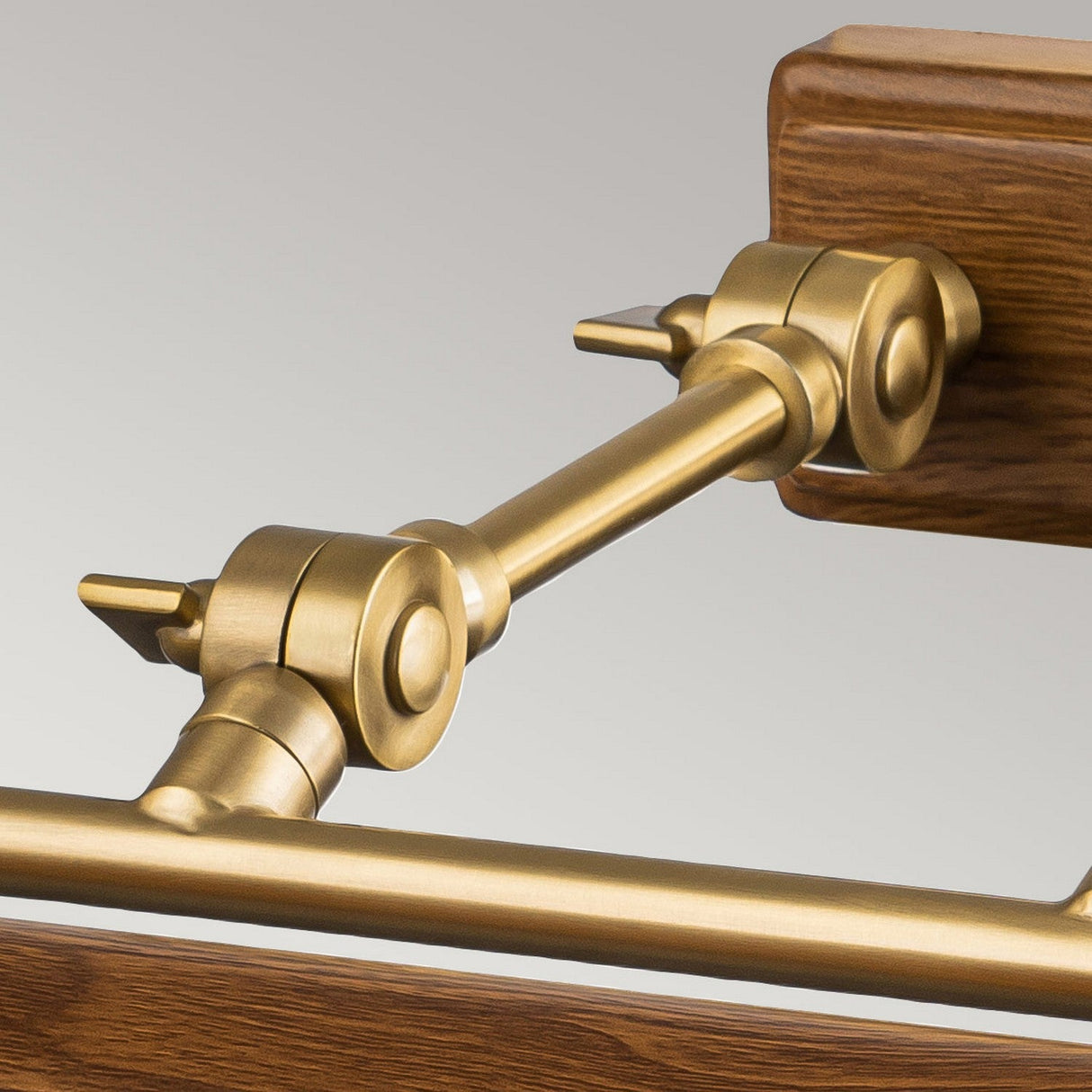 A close-up of the Winchfield 4 Light Large Picture Light in aged brass and oak effect, featuring lever arms against a wooden and metal framework. The polished mechanical joint connects horizontal and vertical rods, highlighting the intricate design and engineering details.
